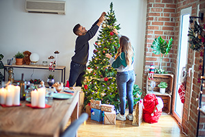 two young people surrounded by holiday decorations help each other place ornaments on a tree and decorate the rest of the house with some happiness and joy and stuff