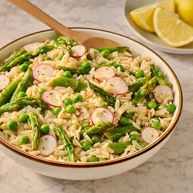 a bowl of orzo pasta with a spoon in the bowl ready for serving sits on the table next to a bowl of lemons. the pasta is topped with peas and asparagus tips and looks very appetizing and the yaller dog sleeps peacefully