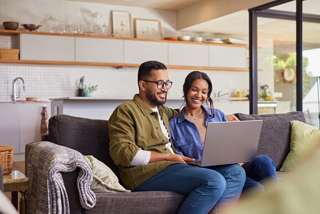 young couple of color smiling on the couch staring at a laptop going over a mortgage application, assessing their chances that a lender will let them have the money they need to own a home and make life comfortable for the yaller dog