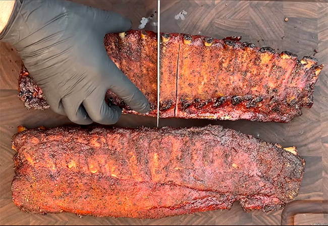 a gloved hand holds down on a rack of ribs seasoned and smoked to perfection on  a wood-grained cutting board as a knife slices up the ribs into individual servings and one for the yellow dog