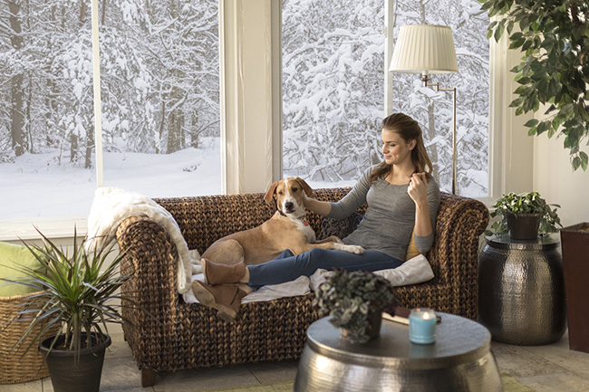 big windows in the room show a snowy wintery scene of cold while a woman seated on a couch near the windows with dog seems warm and comfortable in her warmer older home upgraded and winterized