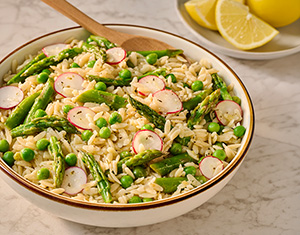 a bowl of orzo pasta with a spoon in the bowl ready for serving sits on the table next to a bowl of lemons. the pasta is topped with peas and asparagus tips and looks very appetizing